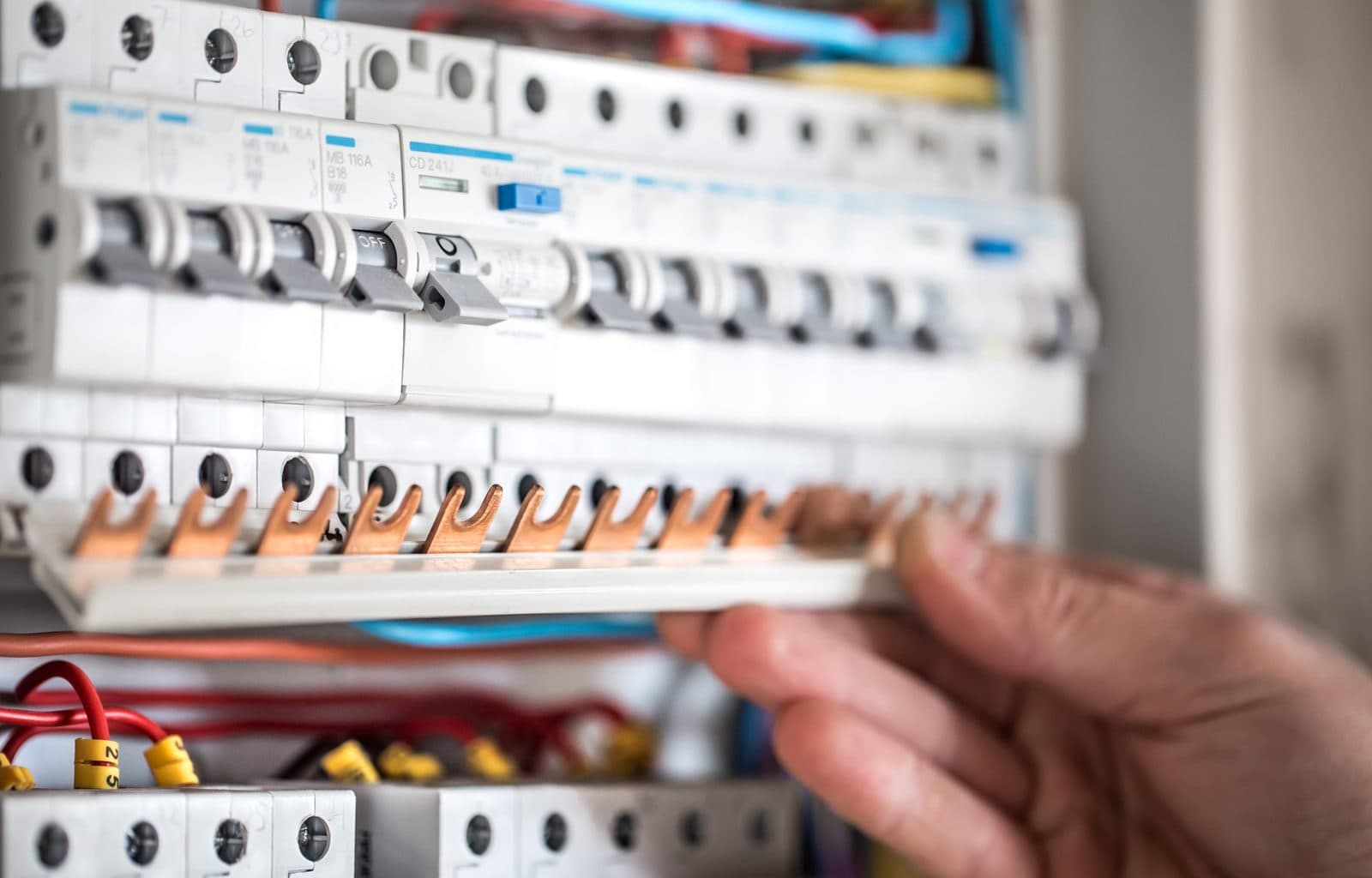 A hand fits a copper busbar into a row of circuit breakers in a fuse box